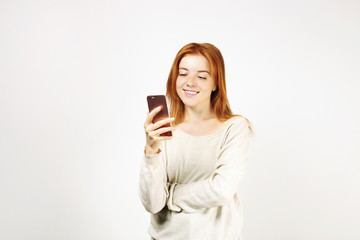 Fototapeta premium Young attractive natural redhead woman smiling at smartphone screen, using mobile phone, wearing loose white shirt. White background, copy space. Beautiful female using online dating app concept.