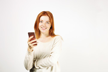 Fototapeta premium Young attractive natural redhead woman smiling at smartphone screen, using mobile phone, wearing loose white shirt. White background, copy space. Beautiful female using online dating app concept.
