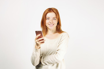 Fototapeta premium Young attractive natural redhead woman smiling at smartphone screen, using mobile phone, wearing loose white shirt. White background, copy space. Beautiful female using online dating app concept.