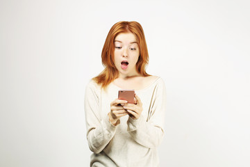 Surprised natural redhead woman with open mouth wearing white oversized loose shirt staring at cell phone screen. amazed hipster ginger female w/ smartphone. Isolated background, copy space, portrait.