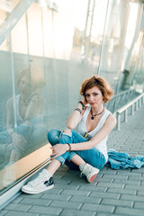 Portrait of attractive woman with short haircut wearing blue jeans posing at the camera while sitting on the floor near the office center window. Outdoors
