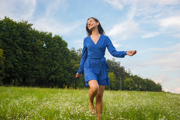 Beautiful smiling young woman runs in a blue dress in a meadow with a flowers at a rural landscape background