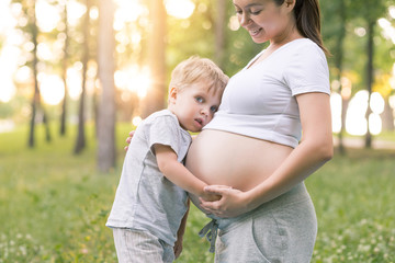 Pregnancy and happy family concept. Little kid boy touching belly of his pregnant mother and smiling at summer nature park