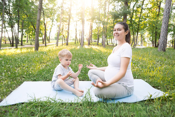 Young beautiful pregnant mother with baby son exercising and doing yoga on blue mat at summer park at sunset. Sportive and healthy motherhood. Fitness, happy maternity and healthy lifestyle concept.