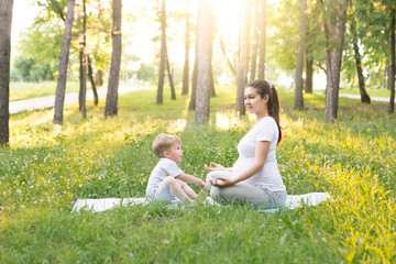 Health and fit motherhood concept. Young beautiful pregnant woman doing fitness exercise in summer park with her little kid son together. FItness materinity outdoors
