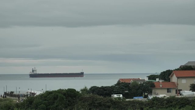 Big industrial barge moving over the horizon of smal countryside town with small red roofs of the building, green hills and sea view of Cantabrian, dramatic mild sky ath the rainy day