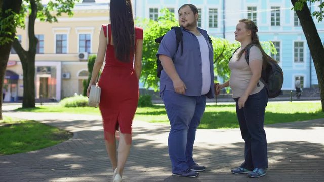 Fat man looking at beautiful lady in red passing by, obese girlfriend jealous