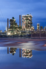 Fototapeta premium Illuminated petrochemical production plant at twilight with dramatic clouds reflected in a pond, Port of Antwerp, Belgium.