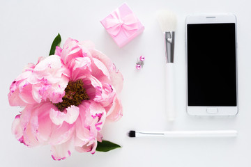 White background with copy space, smart phone lying on the table, pink peony flower, makeup brushes, earrings, small gift for girl, advertising space, top view
