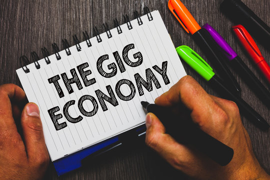 Word Writing Text The Gig Economy. Business Concept For Market Of Short-term Contracts Freelance Work Temporary Hand Holding Pen And Paper Sketch Words Near Lie Some Pen On Woody Desk.