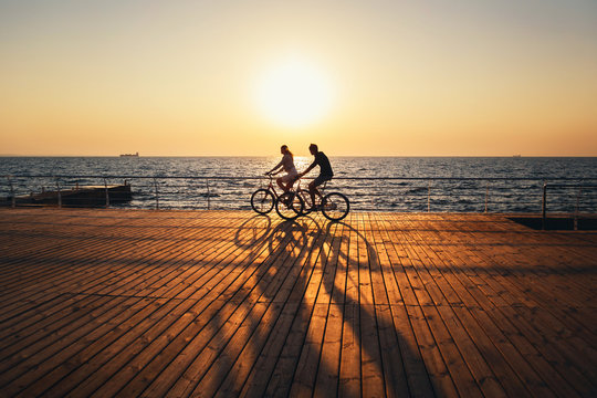 Couple Of Young Hipsters Cycling Together At The Beach At Sunrise Sky At Wooden Deck Summer Time