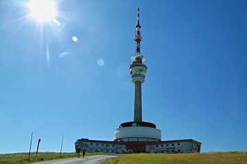 Fototapeta premium Red and white telecommunication and lookout tower Praded, Czech Republic, Europe standing in Jeseniky mountains, backlit with sun, tourists hiking on road, sunny summer day clear blue sky, green trees