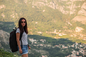 Young beautiful woman with black backpack hitchhiking standing on road. Beautiful young female hitchhiker by the road during vacation trip in mountains. Back view.