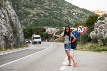 Young beautiful woman with black backpack hitchhiking standing on road. Beautiful young female hitchhiker by the road during vacation trip in mountains. Back view.
