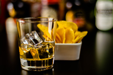 glass of whiskey with ice cubes and salty snacks on the background of the bar