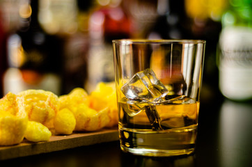 glass of whiskey with ice cubes and salty snacks on the background of the bar