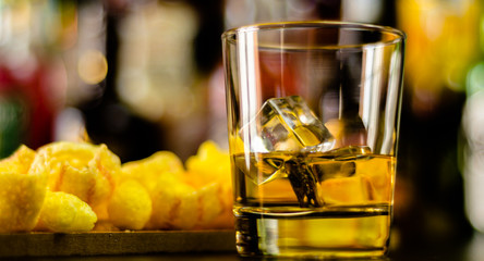glass of whiskey with ice cubes and salty snacks on the background of the bar