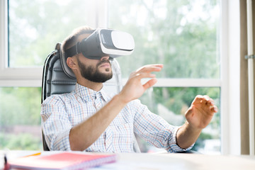 Portrait of handsome bearded man sitting at desk in office and enjoying VR simulator against window in sunlight, copy space