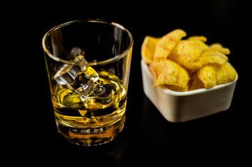glass of whiskey with ice cubes and salty snacks on the background of the bar