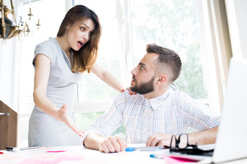 Fototapeta premium Portrait of agitated businesswoman looking shocked while explaining something to handsome colleague sitting at desk in office