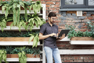 Fototapeta na wymiar Young entrepreneur working outside using modern laptop. Standing near brick wall, plants, eco office. Successful business people.