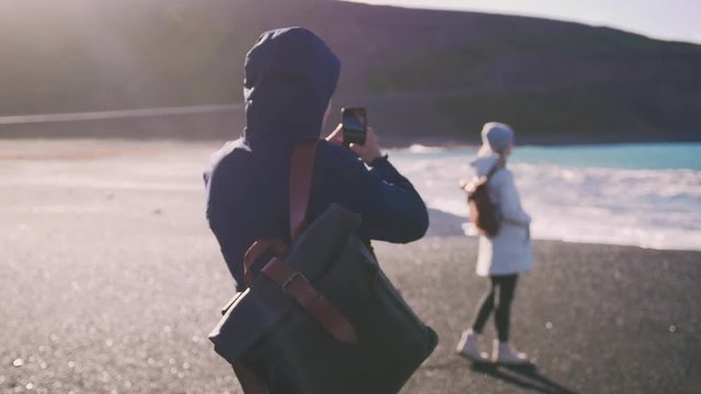 Young loving couple taking pictures on smartphone on beautiful beach in Iceland during sunset, slow motion