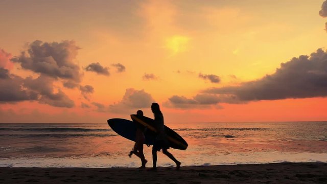 Silhouettes of two surfers walk on beach carrying surfboard against amazing sunset sky and slow motion sea waves rolling on sand beach in Bali island, Indonesia