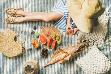 Sommer-Picknick-Einstellung. Frau in gestreiftem Leinenkleid und Strohsonnenhut, die mit einem Glas Roséwein in der Hand, frischem Obst und Baguette auf Decke sitzt, Draufsicht. Outdoor-Treffen oder Lunch-Konzept