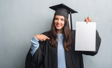 Young brunette woman over grunge grey wall wearing graduate uniform holding degree with surprise...