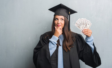 Young brunette woman over grunge grey wall wearing graduate uniform holding dollars serious face...
