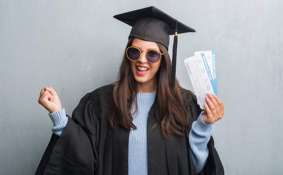 Young brunette woman over grunge grey wall wearing graduate uniform holding boarding pass screaming proud and celebrating victory and success very excited, cheering emotion