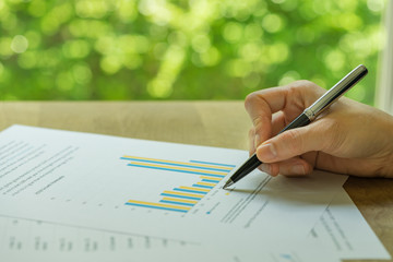 Business, investment or financial report review, hand holding pen reviewing bar graph information data print on paper with green bokeh in the background