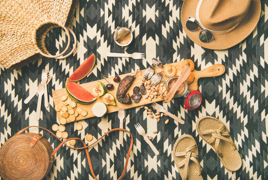 Summer picnic setting. Flat-lay of fresh fruit, smoked sausage, nuts, cheese, pate, cracker on board and woman straw accessories over linen blanket background, top view. Outdoor gathering or lunch