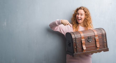 Young redhead woman holding vintage chest very happy pointing with hand and finger - obrazy, fototapety, plakaty