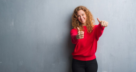 Young redhead woman over grey grunge wall wearing red sweater approving doing positive gesture with hand, thumbs up smiling and happy for success. Looking at the camera, winner gesture.