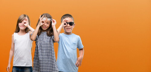 Group of boy and girls kids over orange background with happy face smiling doing ok sign with hand on eye looking through fingers