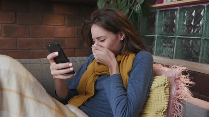 Portrait sick woman lying on the sofa and using her cellphone sneezing virus illness cold ill temperature symptom health close up blanket infection pills medicine slow motion - Powered by Adobe
