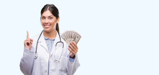 Young hispanic doctor woman holding dollars surprised with an idea or question pointing finger with...