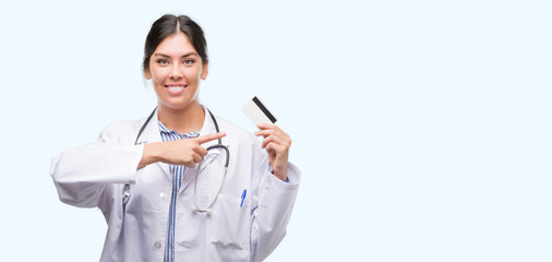 Young hispanic doctor woman holding credit card very happy pointing with hand and finger