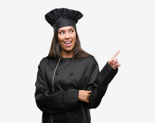 Young hispanic cook woman wearing chef uniform very happy pointing with hand and finger to the side