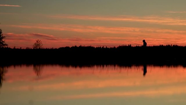 Person standing in a lake fishing during a orange sunset in a forest in the Swedish wilderness