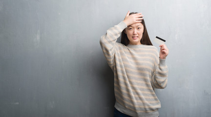 Young Chinese woman over grey wall holding credit card stressed with hand on head, shocked with shame and surprise face, angry and frustrated. Fear and upset for mistake.