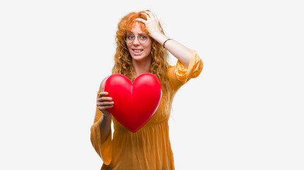Young redhead woman in love holding red heart stressed with hand on head, shocked with shame and surprise face, angry and frustrated. Fear and upset for mistake.