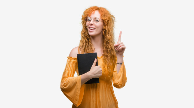 Young redhead student woman holding a book surprised with an idea or question pointing finger with happy face, number one