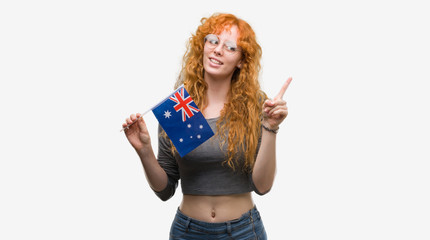 Young redhead woman holding flag of Australia very happy pointing with hand and finger to the side