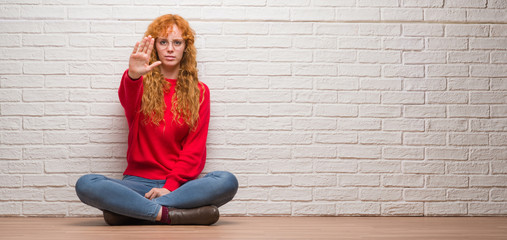 Young redhead woman sitting over brick wall doing stop sing with palm of the hand. Warning expression with negative and serious gesture on the face.