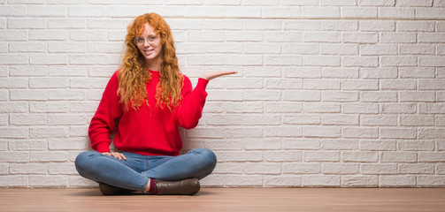Young redhead woman sitting over brick wall smiling cheerful presenting and pointing with palm of...