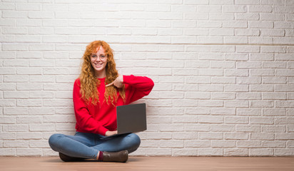 Young redhead woman sitting over brick wall using computer laptop with surprise face pointing...