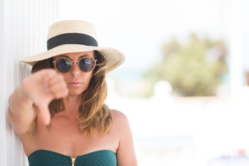 Middle age brunette woman wearing sunglasses and summer hat with angry face, negative sign showing...