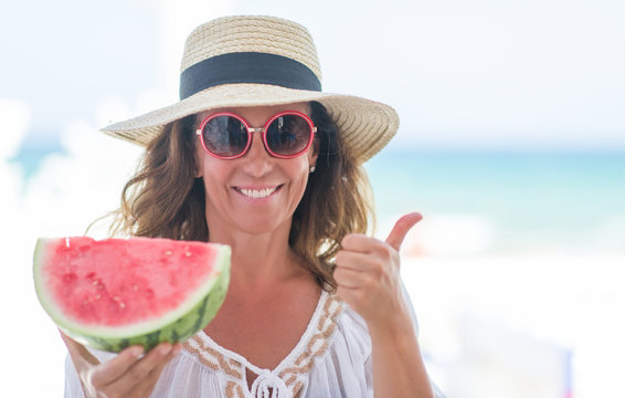 Middle age brunette woman by the sea eating watermelon happy with big smile doing ok sign, thumb up with fingers, excellent sign
